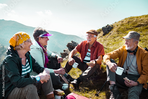 Group of senior friends sitting on mountain hill after long walk. Elderly hikers taking break from hike having drinks and snacks photo