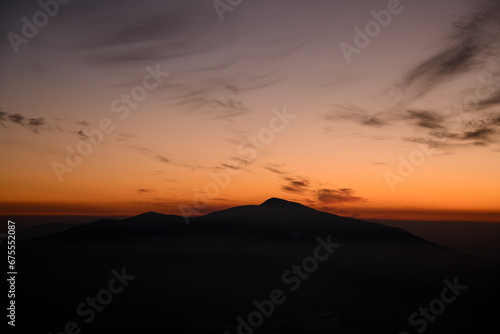 Dark silhouette of mountain against bright orange sunset sky with weightless clouds
