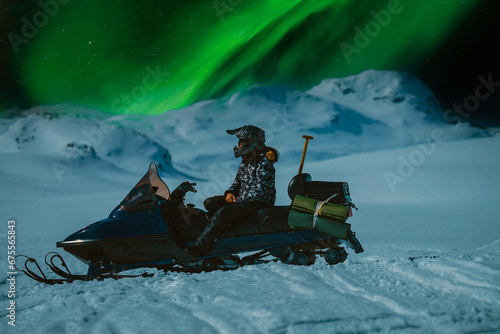Scenic close up side photo of girl sitting on snow mobile with night mountains and northern green lights in starry sky. Driver wear snowmobile helmet and warm overall. Norway, Sweden destinations