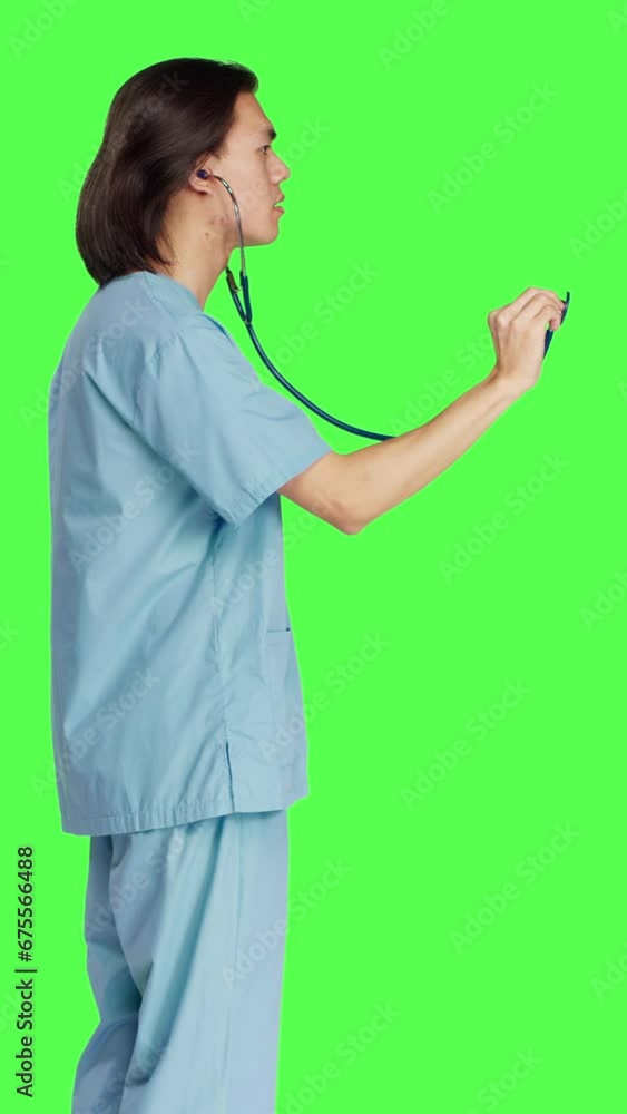 Profile of medic preparing for wellness examination at clinic using stethoscope and wearing medical attire. Asian healthcare assistant holding checkup tool against greenscreen background. Profile of medic preparing for wellness examination at clinic using stethoscope and wearing medical attire. Asian healthcare assistant holding checkup tool against greenscreen background.
