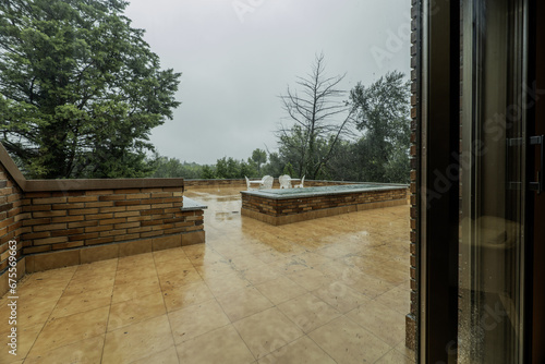 Terrace of a residential home with a white coffee table and matching metal chairs, trees and a lot of rain falling from the sky © Toyakisfoto.photos