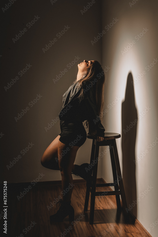 Beautiful young Caucasian woman standing leaning against a wooden stool, wearing black clothing. Beautiful young Caucasian woman standing leaning against a wooden stool, wearing black clothing.