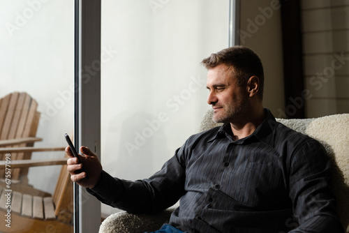 Relaxing businessman at home browsing phone. Man wearing black shirt using smartphone indoors. Happy dad review photos his daughter on vacation. Alone male reading news in his device