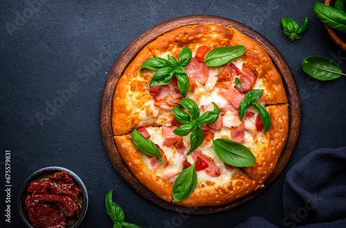 Homemade hot pizza with smoked ham, cheese, spicy tomato sauce and green basil on black table background, top view