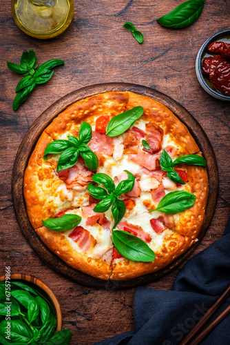 Homemade pizza with pancetta, mozzarella cheese, spicy sauce and green basil leaves on old wooden table background, top view