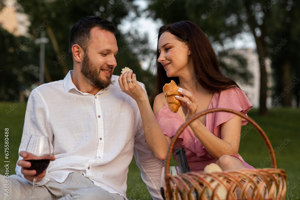Fototapeta premium Cute young girlfriend feeds her boyfriend french yummy croissant on picnic. Couple having romantic date outdoors. Wife giving husband a small piece of bakery in park. Family vacation