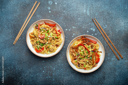 Two bowls with Chow Mein or Lo Mein, traditional Chinese stir fry noodles with meat and vegetables, served with chopsticks top view on rustic blue concrete background.