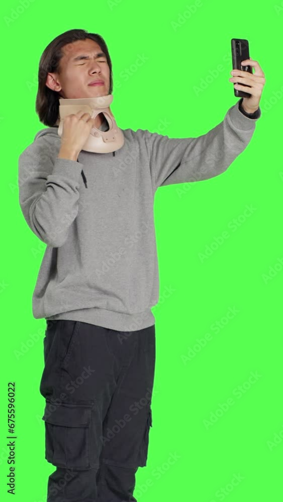 Side view of injured man with cervical collar on videocall against greenscreen background, talking on online web conference while he wears medical neck brace after a fracture. Young guy with cervical Side view of injured man with cervical collar on videocall against greenscreen background, talking on online web conference while he wears medical neck brace after a fracture. Young guy with cervical