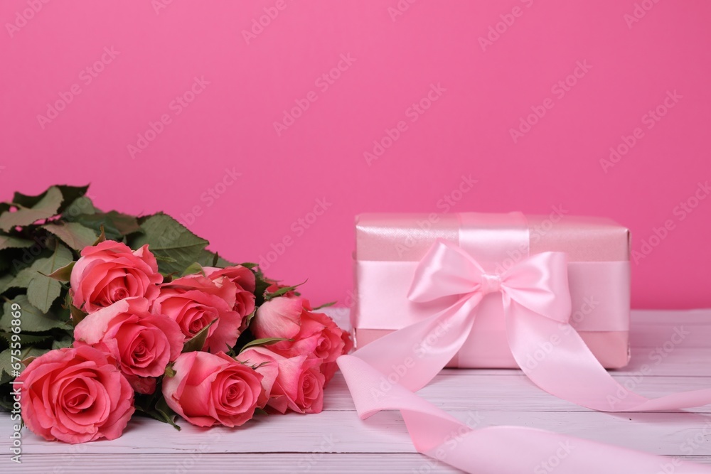 Gift box with bow and bouquet of beautiful roses on white wooden table against pink background, space for text Gift box with bow and bouquet of beautiful roses on white wooden table against pink background, space for text