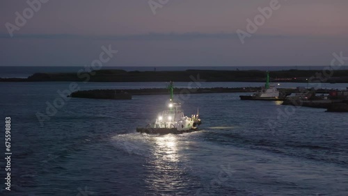 Tug boat with bright lights by breakwater at harbor entrance at sunset