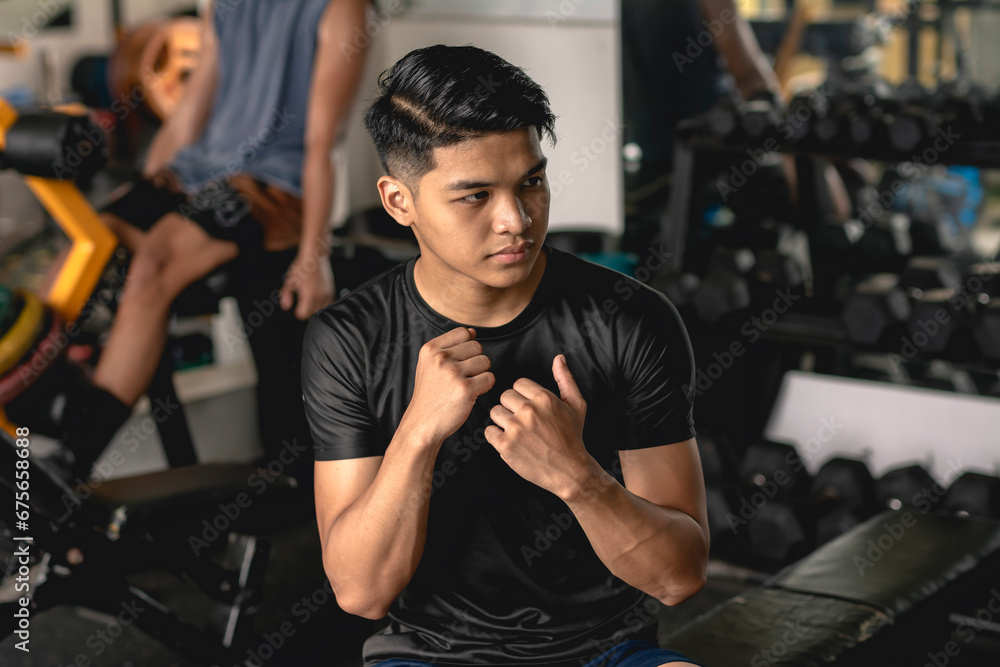 A young handsome asian man practicing his boxing combo skills while sitting on a bench at the gym. Muscle memory and reflex example. - obrazy, fototapety, plakaty 