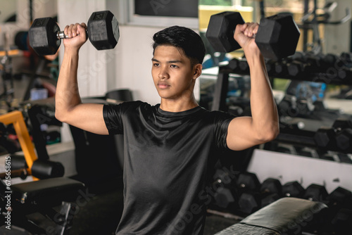 A young asian man in a black moisture-wicking polyester shirt doing a set of seated dumbbell presses at the gym. Shoulder workout and training. photo