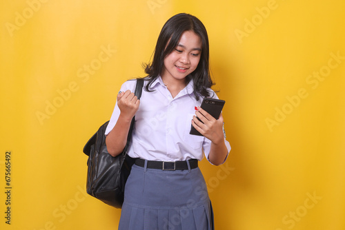 Happy Asian tudent clenching fist showing victory gesture while looking at smartphone and wearing high school uniform and backpack photo