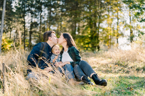 Dad and mom kiss while sitting with a little girl in a clearing near the forest