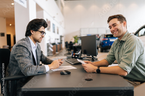 Cheerful male customer talking about terms of buying car with dealer wearing business suit sitting at desk in auto dealership. Concept of choosing, buying new auto at showroom.