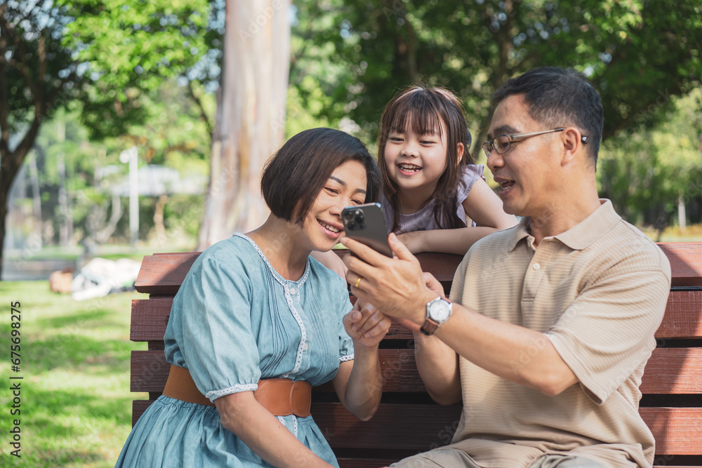 Cheerful Asian family of three having fun watching a phone together in the summer outdoors at the park. Cheerful Asian family of three having fun watching a phone together in the summer outdoors at the park.