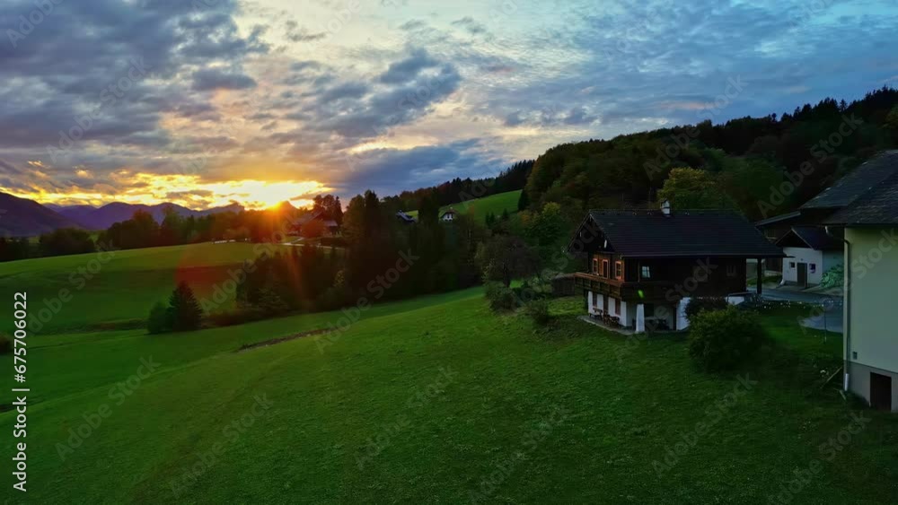 Flying along the green landscape near houses and trees with sun setting behind the mountain summits in the far distance Flying along the green landscape near houses and trees with sun setting behind the mountain summits in the far distance