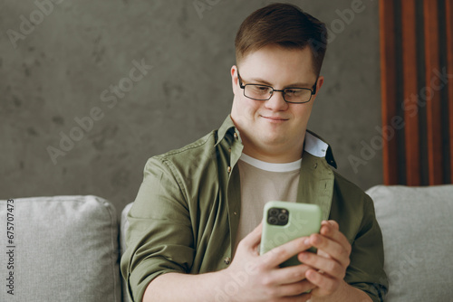 Young cool man with down syndrome wears glasses casual clothes use mobile cell phone sits on grey sofa couch stay at home flat rest spend free time in living room. Genetic disease world day concept.