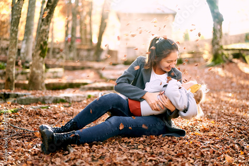 Smiling mom hugging little girl on her knees while sitting in autumn park under falling leaves photo