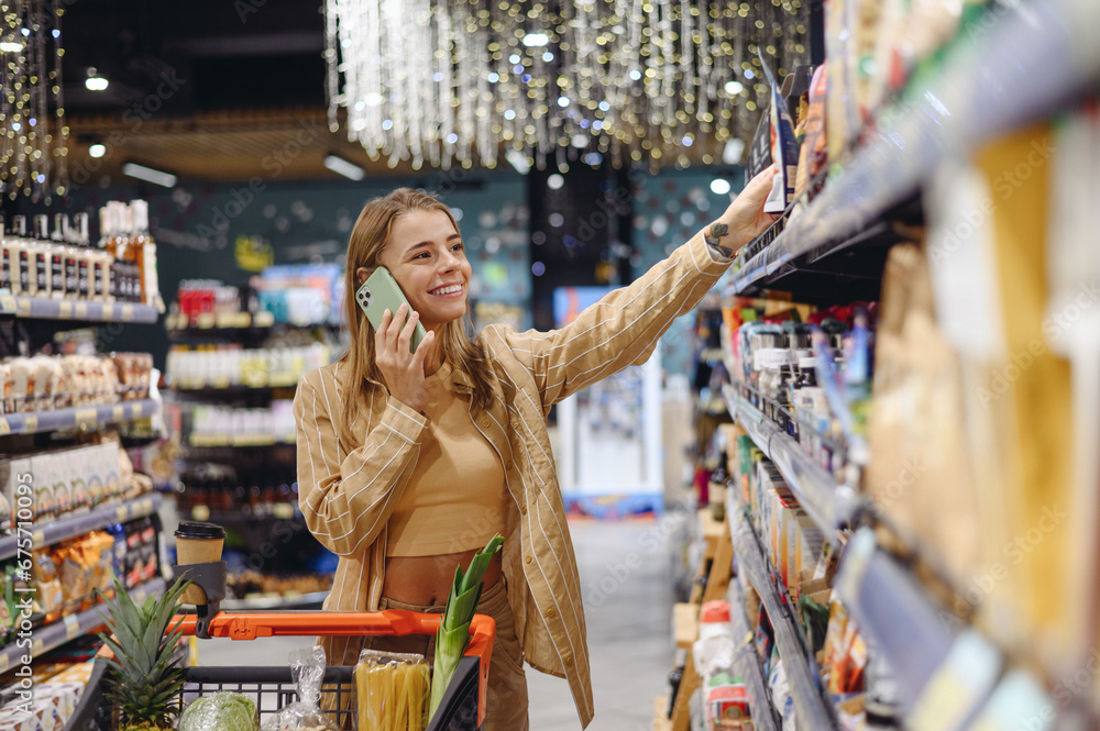 Young customer woman wear casual clothes talk on mobile cell phone shopping at supermaket store grocery shop buy with trolley cart choose products in hypermarket. Purchasing food gastronomy concept.