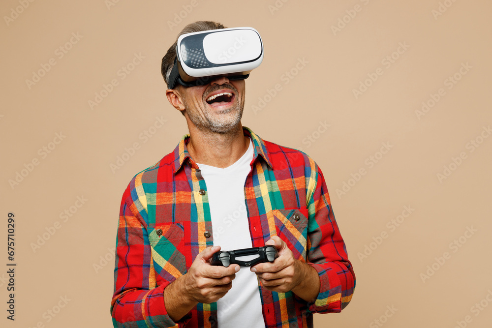 Fototapeta premium Adult man wear red shirt white t-shirt casual clothes watching in vr headset pc gadget hold play pc game with joystick console isolated on plain pastel light beige color background. Lifestyle concept.