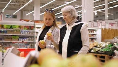 An elderly lady and her adult granddaughter are buying groceries at the supermarket, they are sniffing fresh apples. An old lady with a woman in a grocery store. photo