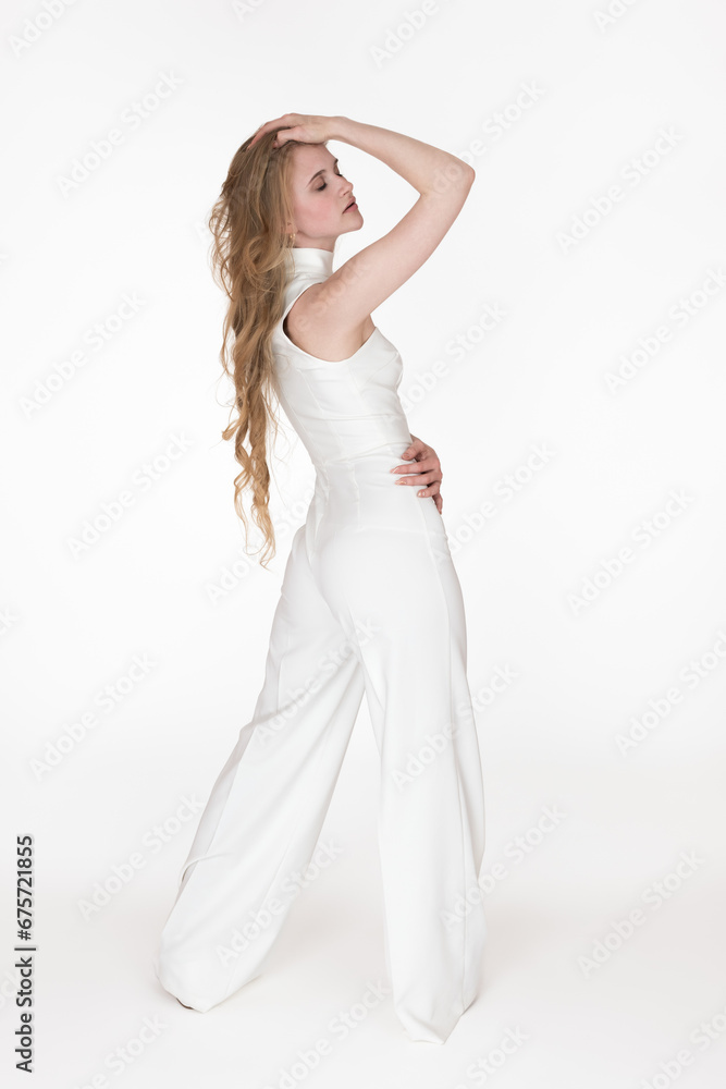 Back view of woman with eyes closed and one hand on head, other on waist. Full length view of Caucasian blondie woman wearing white long jumpsuit posing, standing on white background. Studio shot