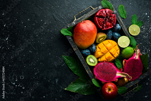 Tropical fruits in a wooden box: mango, dragon fruit, lime, pomegranate, plum, apple. On a black stone background.