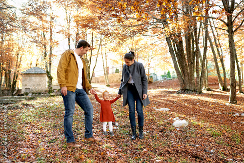 Mom and dad look at a little girl, holding her hands in the autumn forest