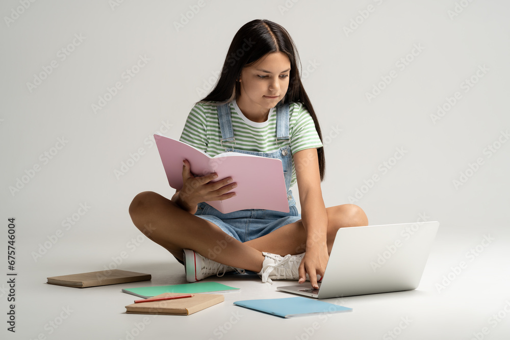 Intelligent teenage girl sitting on floor with textbook in hands, doing homework, thinking, reading article from laptop computer. Books, textbooks, pens, school articles lying disorderly on floor Intelligent teenage girl sitting on floor with textbook in hands, doing homework, thinking, reading article from laptop computer. Books, textbooks, pens, school articles lying disorderly on floor