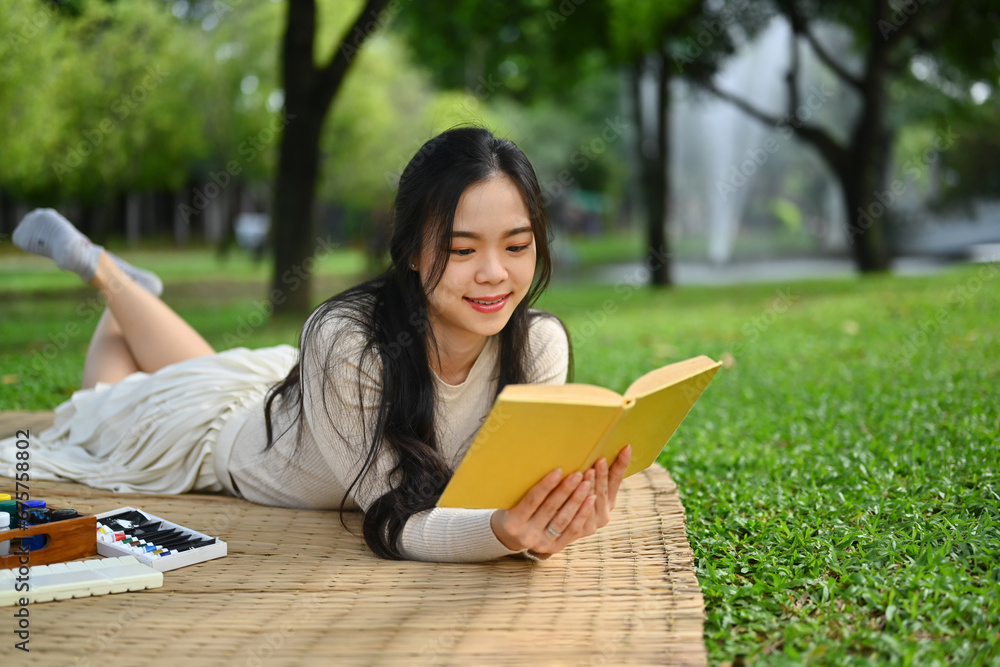 Obraz premium Relaxed young woman lying down on mat and reading interesting book at green park