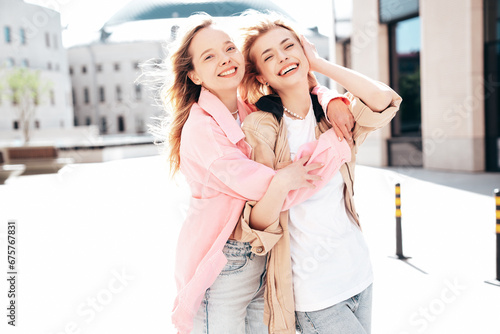 Two young beautiful smiling hipster female in trendy summer clothes. Carefree women posing in the street. Positive models having fun outdoors at sunny day. Cheerful and happy. Wind blows hair