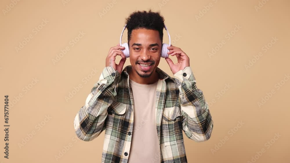 Young man of African American ethnicity he wears shirt casual clothes listen mp3 music in headphones slow dance sing song have fun enjoy relax show thumb up isolated on plain beige brown background Young man of African American ethnicity he wears shirt casual clothes listen mp3 music in headphones slow dance sing song have fun enjoy relax show thumb up isolated on plain beige brown background