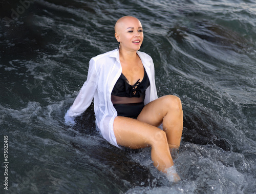 beautiful woman with alopecia relaxing on seashore sitting or dragging chair in water sunset or sunrise time. girl with no hair is walking on beach dancing with white shirt as with a man. crazy fun ti