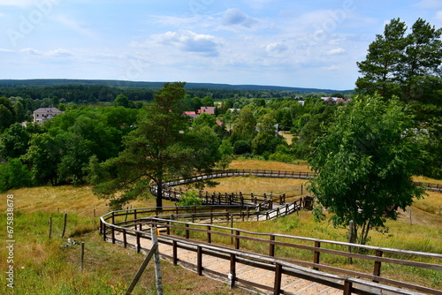 A view from a top of a tall hill or mountain covered with grass and trees showing some small village or cottage in the distance, wooden path with handles leading upwards and observation spot