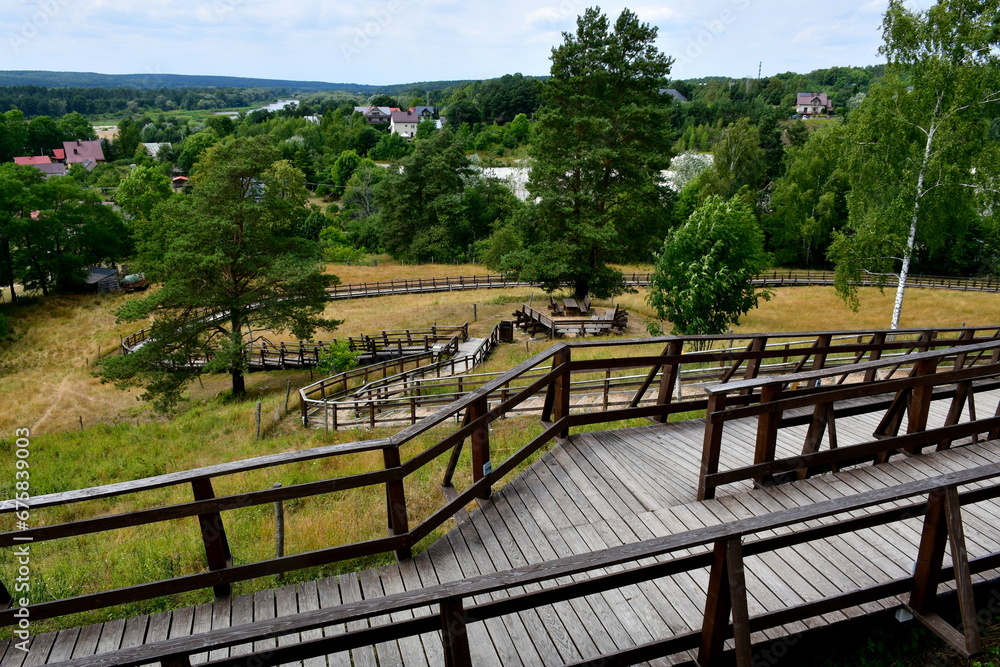 A view from a top of a tall hill or mountain covered with grass and trees showing some small village or cottage in the distance, wooden path with handles leading upwards and observation spot