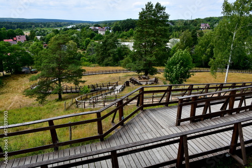 A view from a top of a tall hill or mountain covered with grass and trees showing some small village or cottage in the distance, wooden path with handles leading upwards and observation spot