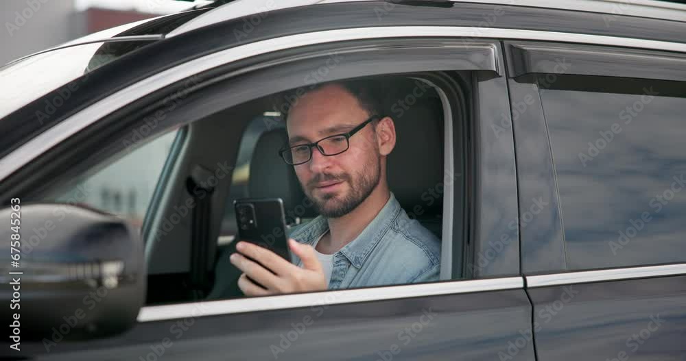 Young Caucasian man sits in a car on a parking lot and reading from a smart phone Young Caucasian man sits in a car on a parking lot and reading from a smart phone