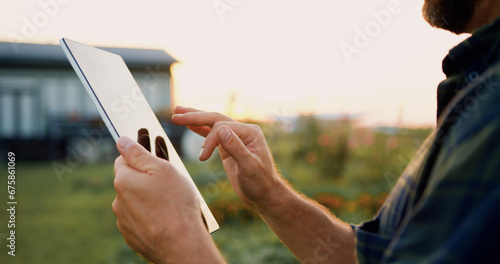 Businessman promptly carries out online deal to sell crop. Close up of farmers hands using digital computer and searches for information in tablet in agricultural field with plants in windy weather on