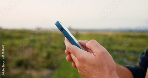 Close up of farmers hands taking notes and searches for information in smartphone in vegetables field with plants in windy weather on sunset. Businessman promptly carries out online deal to sell crop.