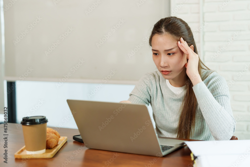 Beautiful Asian business woman in gray sweater sitting working in front of laptop, looking stressed, holding hands on emples. Next to it croissant brown disposable coffee cup. Insmall business cafe Beautiful Asian business woman in gray sweater sitting working in front of laptop, looking stressed, holding hands on emples. Next to it croissant brown disposable coffee cup. Insmall business cafe
