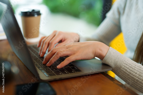 The hands of a young Asian businesswoman, a cafe owner, are typing a coffee product income summary into a laptop, next to which are disposable paper coffee cups. small business cafe