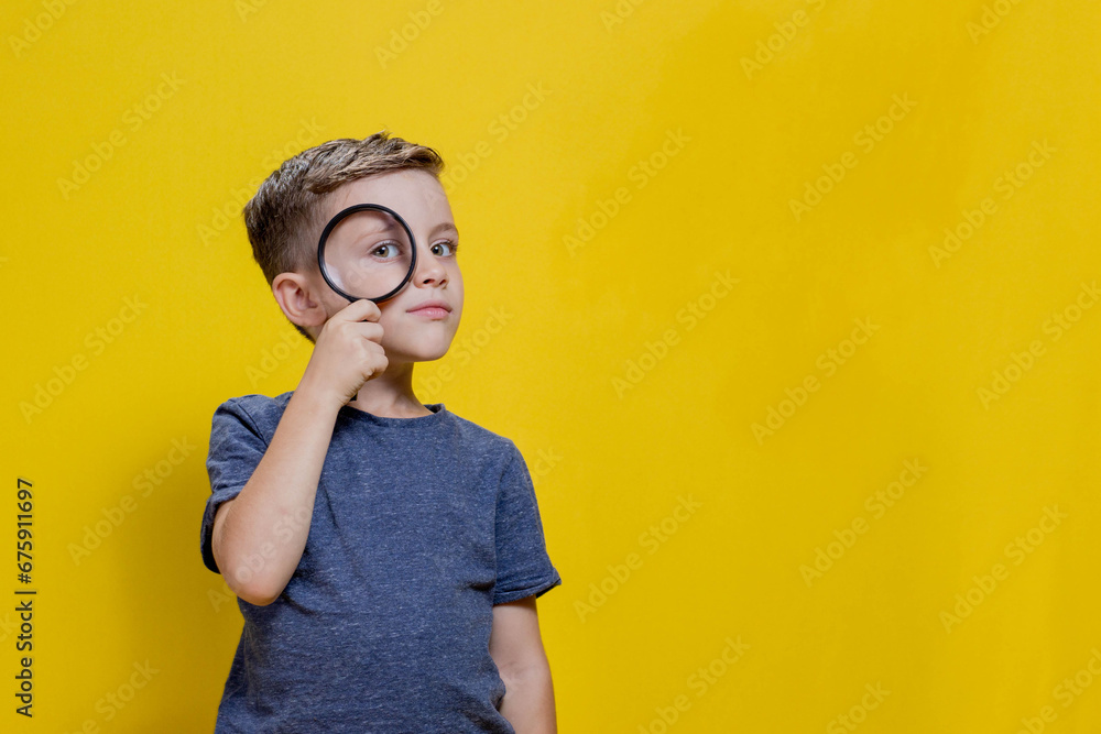 Positive curious schoolboy in casual clothes looking at camera through magnifying glass while standing on yellow background. Copy space - obrazy, fototapety, plakaty 