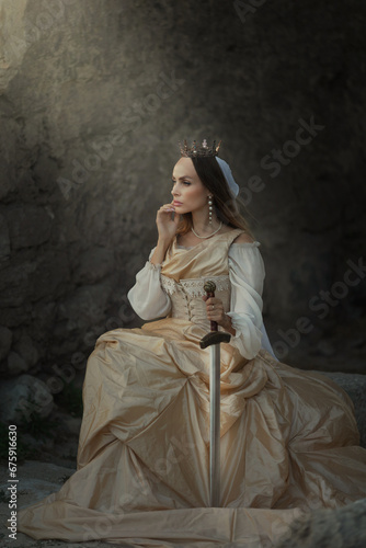 Fotografie A beautiful red-haired girl in an old medieval dress with swords stands in the mountains and looks into the distance