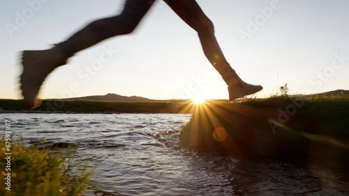 Two traveler in trekking boots run and jump through wild water of small river at sunset. Active sport recreation, adventure in hike and tourist way
