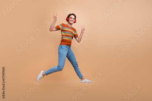Full length portrait of energetic excited person jump show v-sign listen music headphones empty space isolated on beige color background