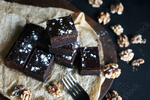 Chocolate brownie next to walnuts and a black fork on a wooden board on a dark background © Kateryna
