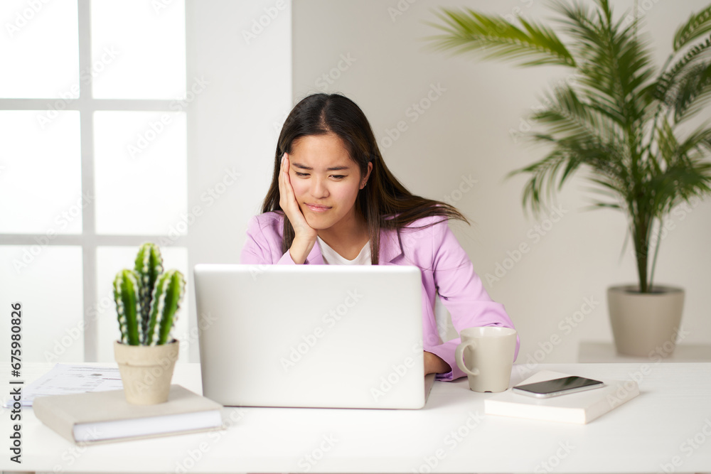 Asian businesswoman resting her hand on her chin and looking at computer screen in office Asian businesswoman resting her hand on her chin and looking at computer screen in office