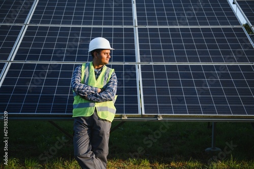 Portrait of Young indian man technician wearing white hard hat standing near solar panels against blue sky. Industrial worker solar system installation