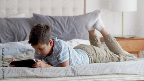 A little boy lies on stomach and moves his feet on the sofa in the bedroom in positive mood. A child watches a funny movie or cartoon, using a tablet, smiles and laughs. The concept of happy and photo